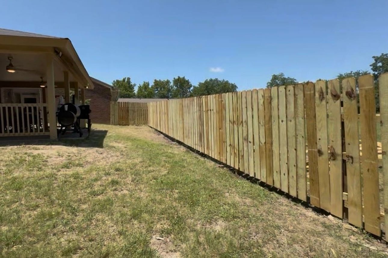A newly built wooden dog-ear privacy fence running the length of a back yard, with a gate, beside a covered patio